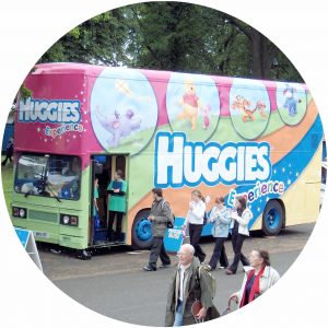 The Huggies promotional roadshow bus at the Higland Showground. THis has all the windows of the bus covered over with panelling overwhich is a very colourful branding featuring cartoon characters using pastel colours. Two promotional staff are seen standing in the bus doorway with people walking past.