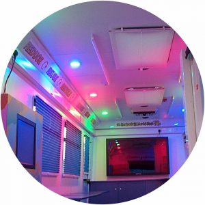 A vibrantly illuminated interior using multi coloured lighting for a childrens playbus. The walls are white but the lighting throws many colours off them to create a disco type feel. Windows are fitted with venetian blinds for privacy.