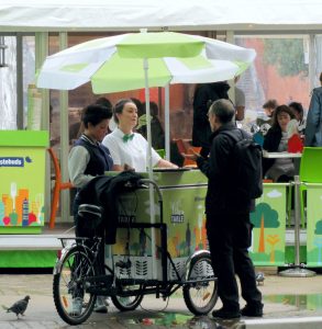 A photo of an ice cream bicycle with umbrella. A sales person is talking to a customer to whome they are about to service some ice cream