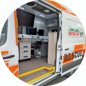 Looking inside of a search and rescue van via the side door. Inside can be seen a worktop with screens and pc monitors. There is also a small filing cabinet In the ceiling is an air conditioning unit.