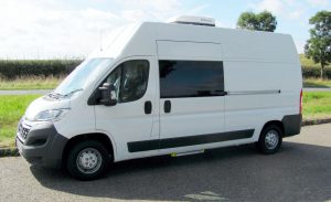 Nearside view of a plain white Surveillance van fitted tinted side door window and slide out step visible. A roof mounted air conditioning unit can aslo be seen. The background is of a main road.