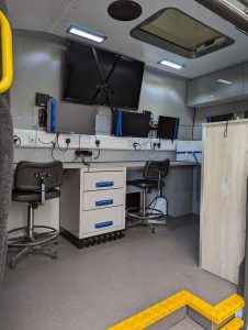 A photo facing through the side door of a surveilland vehicle showing a work top with two seating positions facing three small screens and one large one.