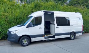 With bushes in the background this is a nearside view of a smaller sized surveillance van. The side door is open to show a cabinet and wall mounted screen.