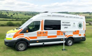 Exterior view of a van converted into an incident command unit. It is white with orange patches with a countryside background.