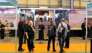 THis is an example of an exhibition vehicle being used as an exhibition stand inside of an exhibition hall. It is a close up and shows open glazed patio doors with steps leading up to them and inside can be seen a range of drinks dispensing machines which are for display purposes. There are people standing in front of the stand.