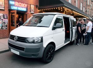 A small camper van conversion designed for a musician that contains space for playing musical instruments. The vehicle is white and located outside of a music venue