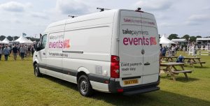 A converted van that has been installed with a large number of charging points for hand held payment terminals used at events. The van is plain white in colour and shown in the background is an outdoor event.