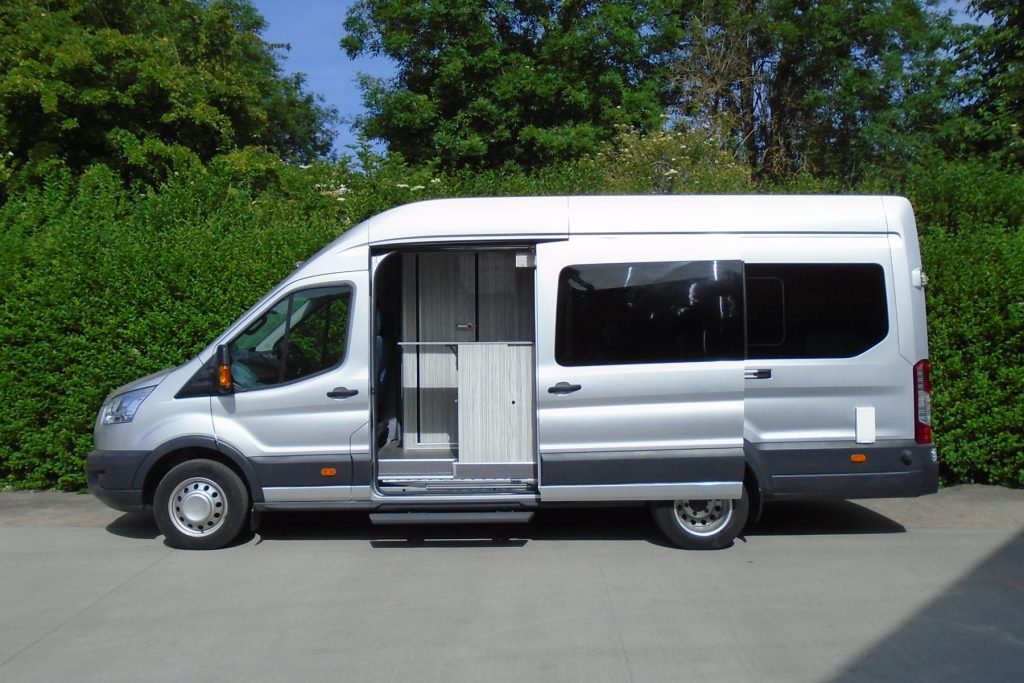 Side view of a camper van conversion showing the side door open, revealing the small galley just inside the door. The van is silver with green bushes in the background