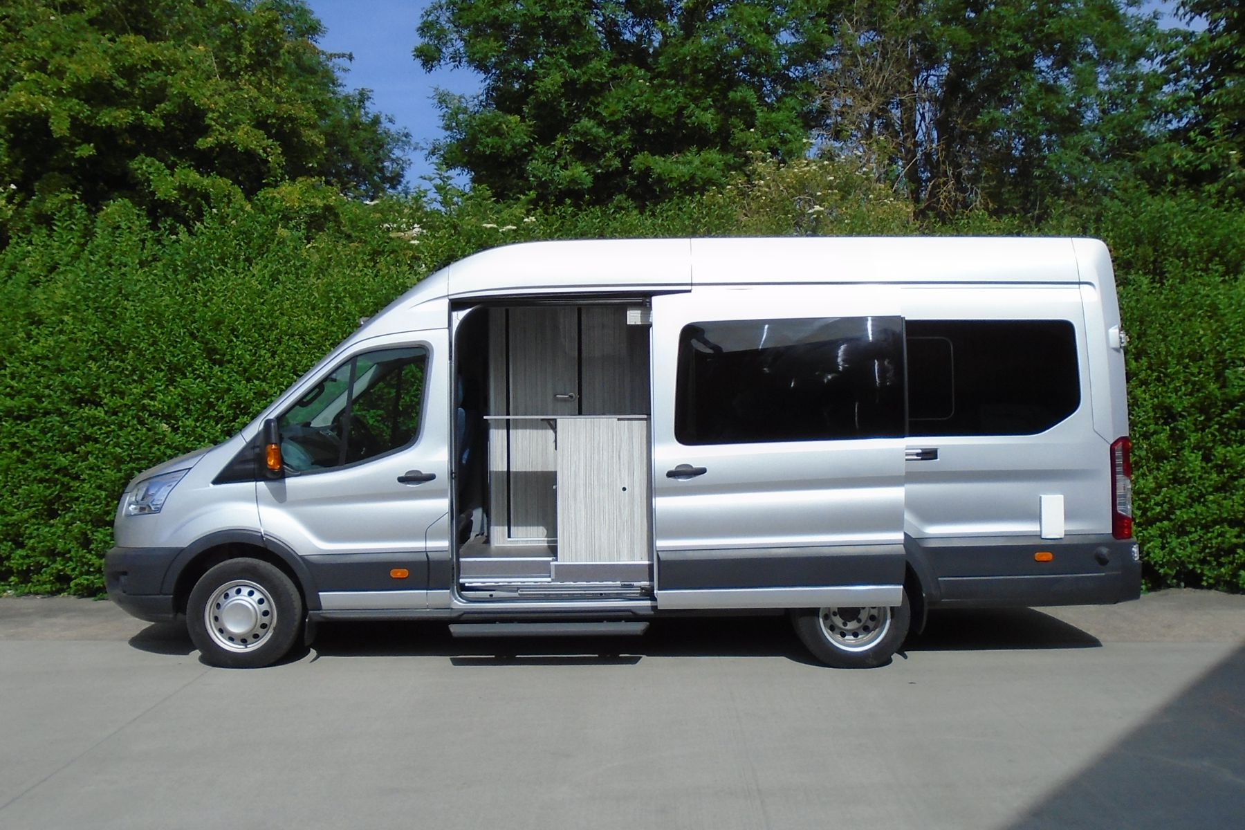 Side view of a camper van conversion showing the side door open, revealing the small galley just inside the door. The van is silver with green bushes in the background