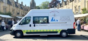 A converted van in which a full mobile veterinary surgery. has been installed. The van is white with a window showing in the side door. On the roof can be seen a roof-mounted air conditioning unit. The background is of a market square in a Cotswold village