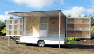 This bespoke trailer is based on a single axle chassis and features a 3metre box body with a hinge up canopy and two swing out panels which each contain shelving similar to the interior of the trailer. This small car towable trailer is white in colour with wooden decorative panels behind the shelves. It was designed to be used as a small retailing unit selling pet dog equipment.
