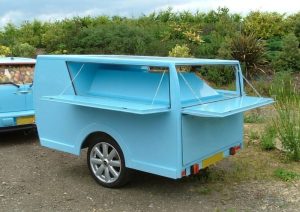 An unusual single-axle small trailer painted blue. It is attached to a mini car and is no higher than it. Three small shelves fold down to reveal a product display area. This is designed to provide a base for product sampling promotional activities.
