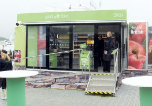 Bespoke trailers are often used for mobile retailing, and in this photo, we show a 7 metre trailer with entrance steps and a wheelchair ramp leading to an entrance door, which is open with a shop assistant standing in the entrance. The side of the trailer is fully glazed, and inside can be seen grocery shop shelving packed with snadwiches and drinks