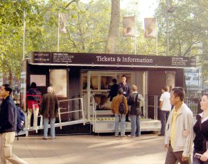 A custom-built mobile ticket office all in black livery, photographed in Leicester Square, London. It features access steps and a wheelchair ramp leading to open glazed patio doors. The banner board reads "Tickets and information"
