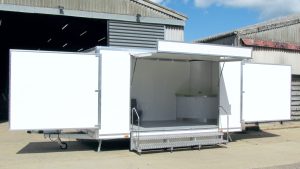 A 5 metre custom-built exhibition trailer featuring a hinge-up canopy with banner board and showing two large swing-out display panels. Steps provide access to the trailer with a curved display counter showing in one corner