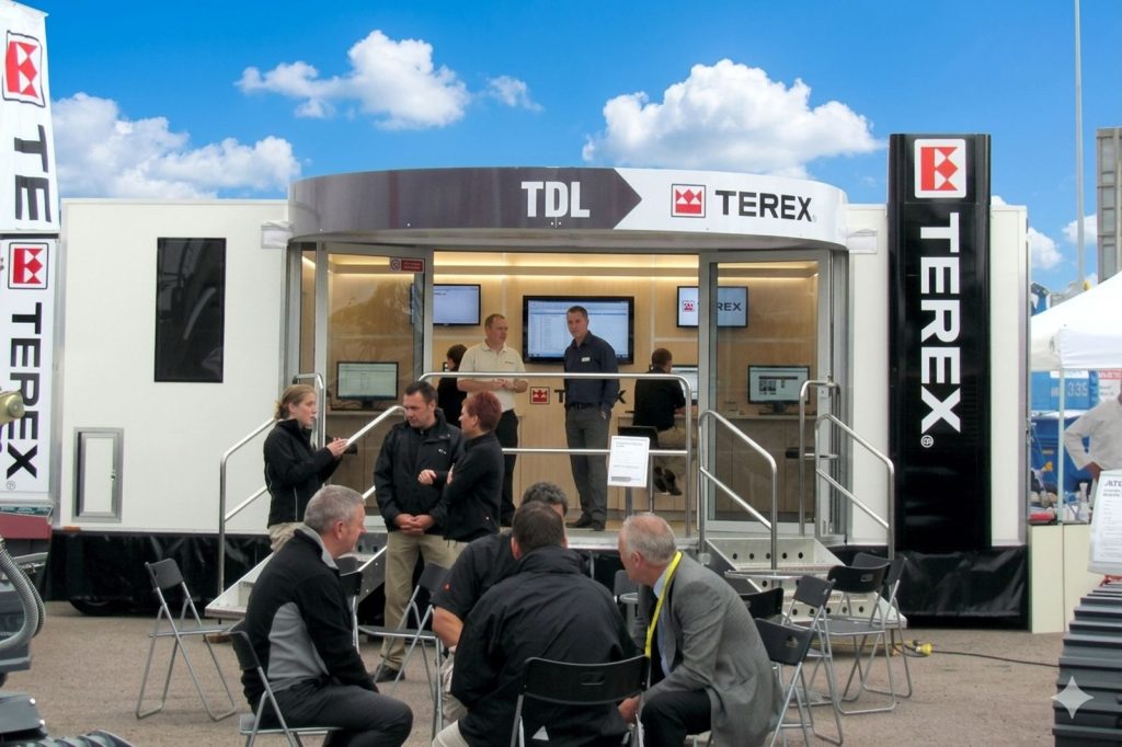 A 7 metre long custom-built exhibition trailer featuring a semi-circular stage and canopy entrance leading to patio doors. Two sets of steps provide access to the platform. There are four people sitting at a table in front of the trailer platform with three people standing behind them. Inside of the trailer can be seen wall mounted screens on a back wall.