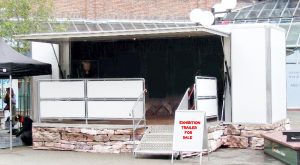 This is one of our 7 metre used exhibition trailers, which we currently have for sale. The body is all in white. A full-length canopy covers a hinge down platform with handrails to create an open and inviting display within the trailer. A set of aluminium steps leads onto the platform. There is a sign in front of the trailer saying "exhibition trailer for sale".