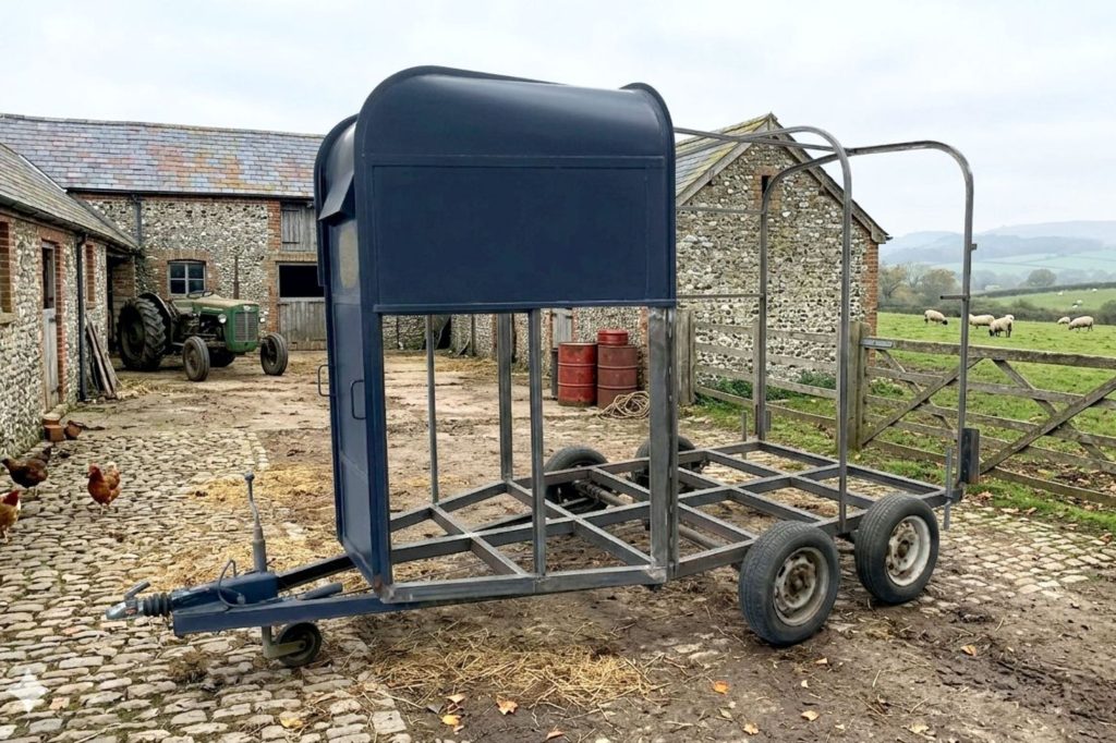 A derelict-looking. old horsebox trailer with sides removed and only rusty steelwork showing. It is located in a farmyard. Even the wheels are all rusted and it looks ready fto be made into scrap.