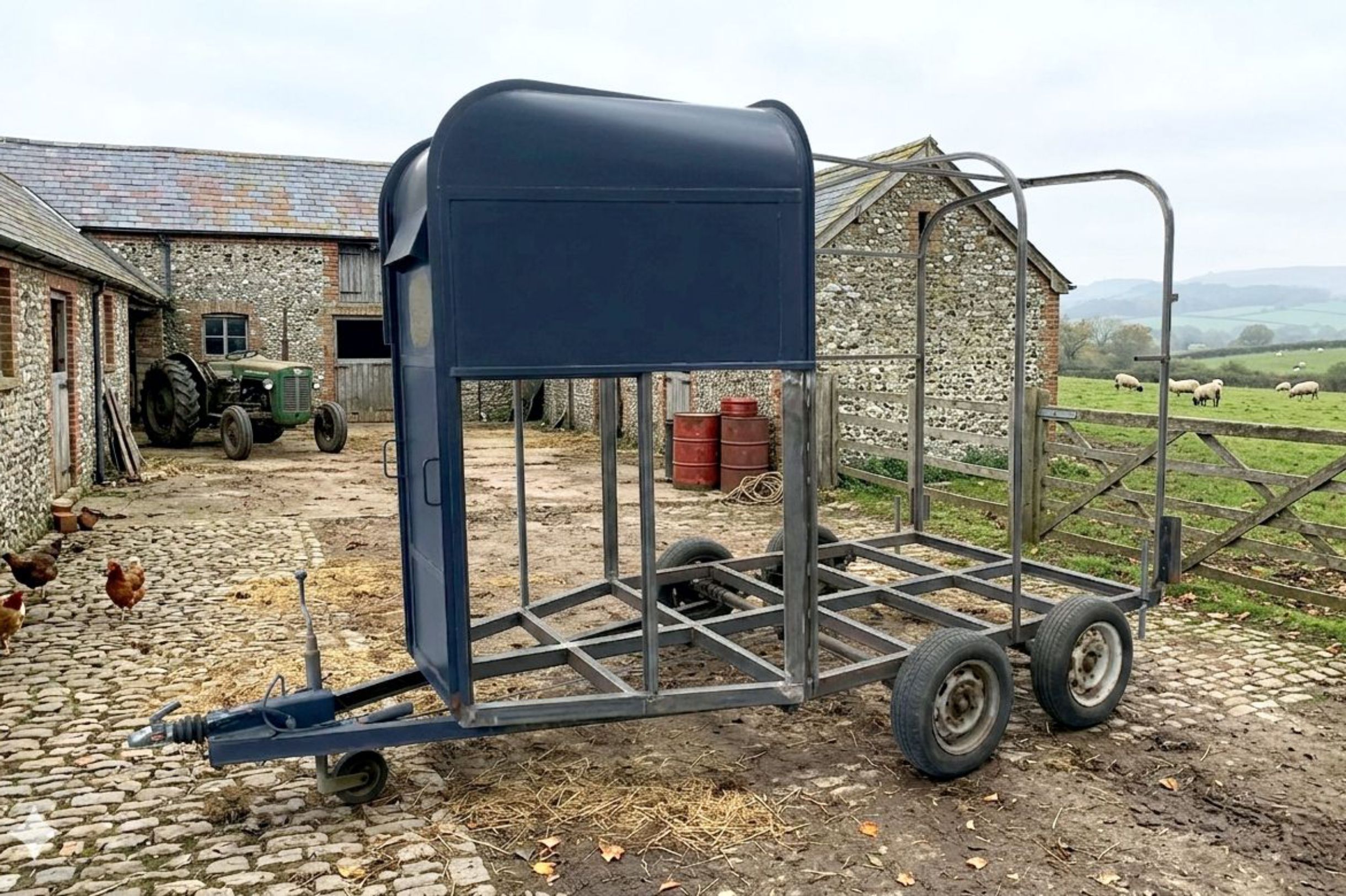 A derelict-looking. old horsebox trailer with sides removed and only rusty steelwork showing. It is located in a farmyard. Even the wheels are all rusted and it looks ready fto be made into scrap.