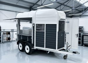 A fully restored horsebox trailer made into a superb catering trailer. It has a bright white roof with charcoal-coloured timber panelling. It has a serving hatch to the side and rear. The trailer is shown in a factory setting, ready for the customer to collect.