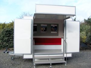 A small 3 metre car towable used exhibition trailer with all graphics removed to show all body panels in bright white. A canopy with a banner board is open to reveal the entrance with seating inside. Two swing out display panels swing to either side of the entrance. Steps provide access into the trailer.