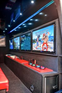 Interior view from the rear door of a gamewagon video gaming van showing seating bench to the left and a row of screens to the right. Be;low the screens is a worktop in red. The rest of the interior is in black.