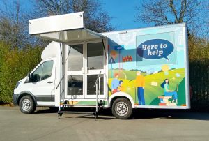 The exterior view of a chassis cab community support vehicle featuring a small hinge up canopy and two glazed entrance doors. signage says"Can we help"