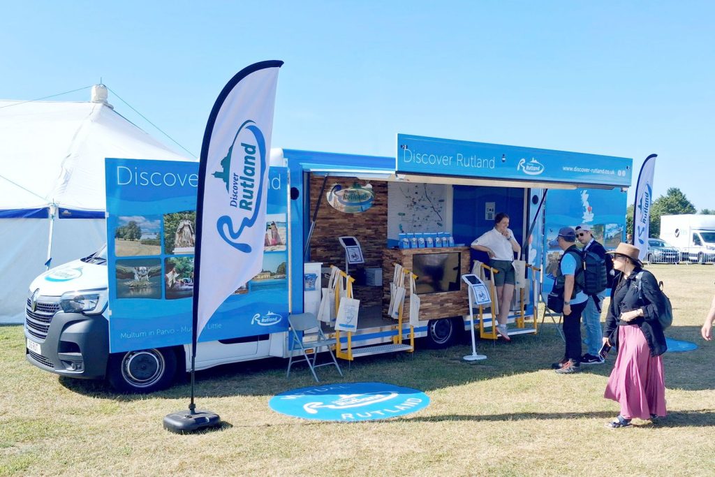 Exterior view of an exhibition vehicle used by Rutland Council to promote "visit Rutland". It is blue in colour and has a wide open frontage for people to see the wall mounted interior dispays.