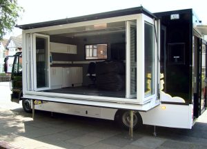 A 7500 kgs chassis cab fitted with an extension pod, in which the main side wall is made up of glazed panels which have been moved to one side to show an open stage-like image is an example of one of our expandable vehicles and trailers